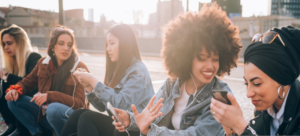 Diverse group of young women sit on a curb talking and looking at each other's phones while smiling and talking