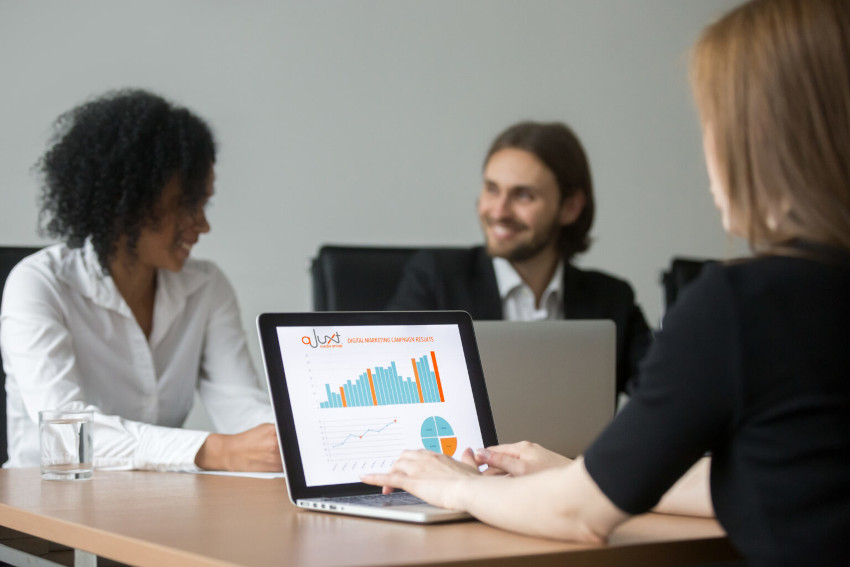 Three young business persons sit at a meeting table with laptops displaying graphs and talking