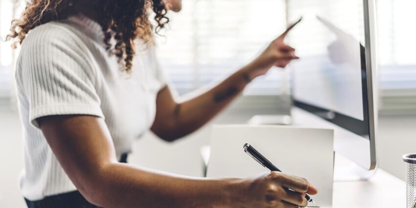 Working from home, a Young adult woman writing in notebook and using touchscreen on computer monitor at a desk