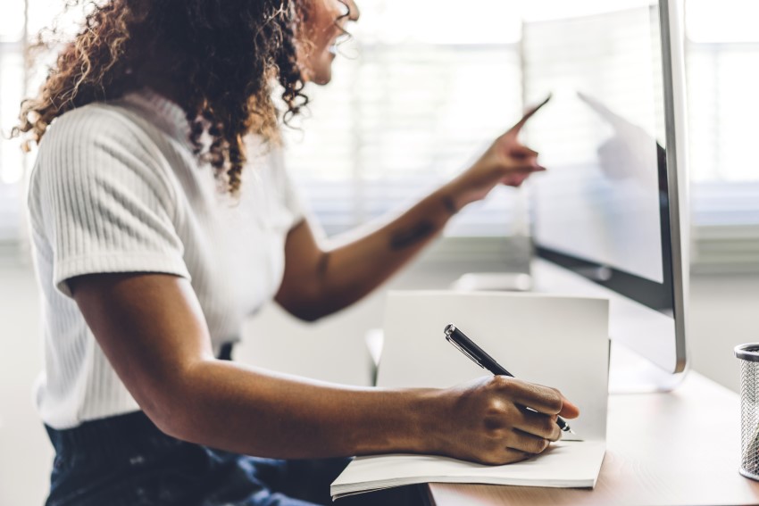 Working from home, a Young adult woman writing in notebook and using touchscreen on computer monitor at a desk