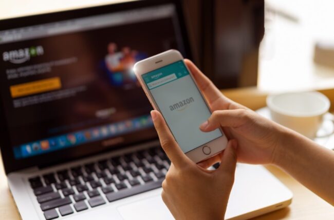 Hand holds smartphone with Amazon logo on screen; in background, laptop with amazon page on screen.