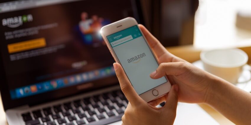Hand holds smartphone with Amazon logo on screen; in background, laptop with amazon page on screen.