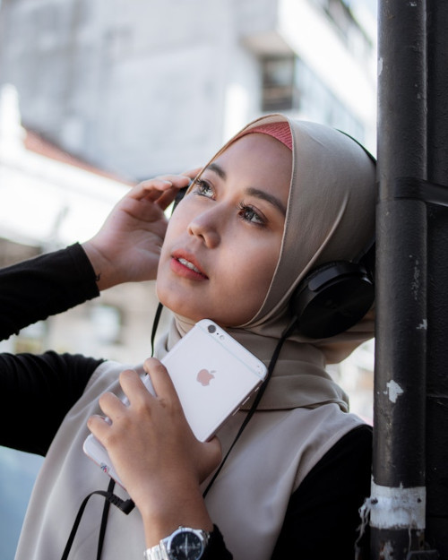 young woman wearing a head scarf and holding an iphone listens to headphones while leaning against a pole outside