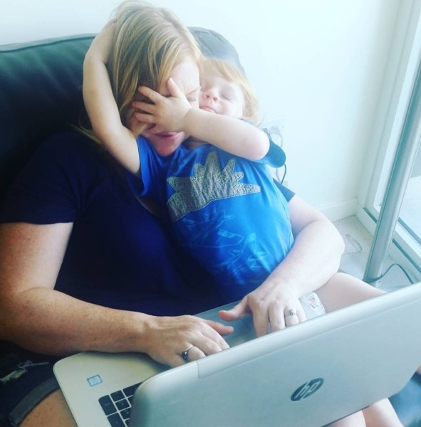 aJuxt CEO Lynsie Slachetka tries to type on her laptop while her toddler covers her face/eyes with his hands while sitting on her lap