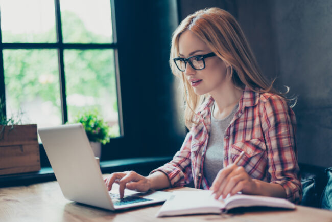 Portrait of busy young person working in workstation on computer searching important information and writing it out in her copybook