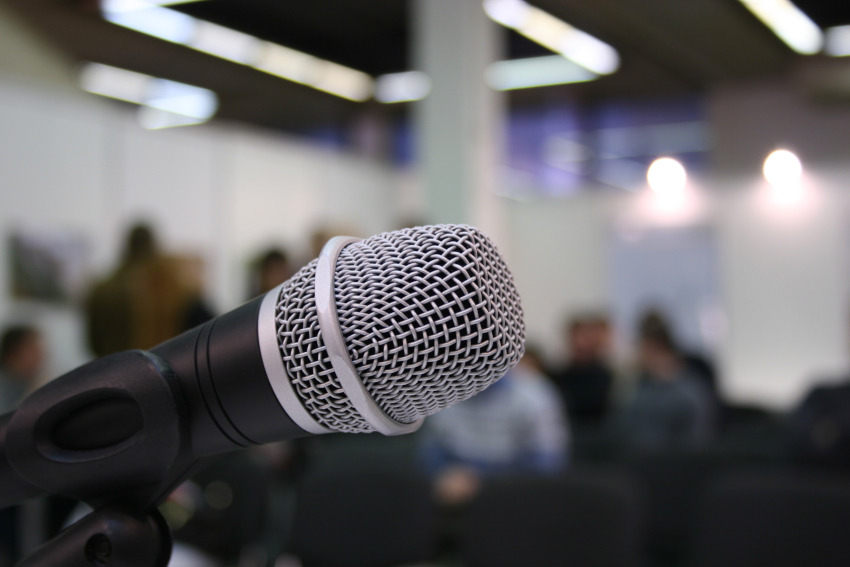 Close-up on microphone in the foreground with a large auditorium with people is in the background.