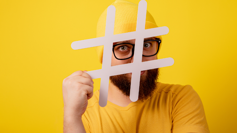 Man with full beard and black-rimmed glasses peeks through an oversized hashtag