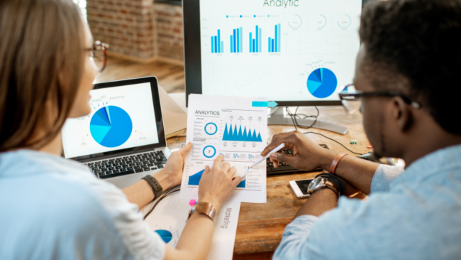 a woman and man talking over multiple and screens covered with a variety of graphs and other data