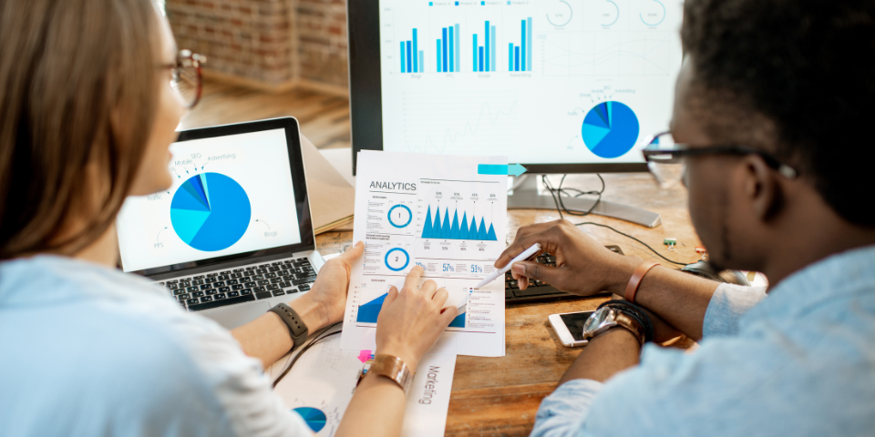 a woman and man talking over multiple and screens covered with a variety of graphs and other data