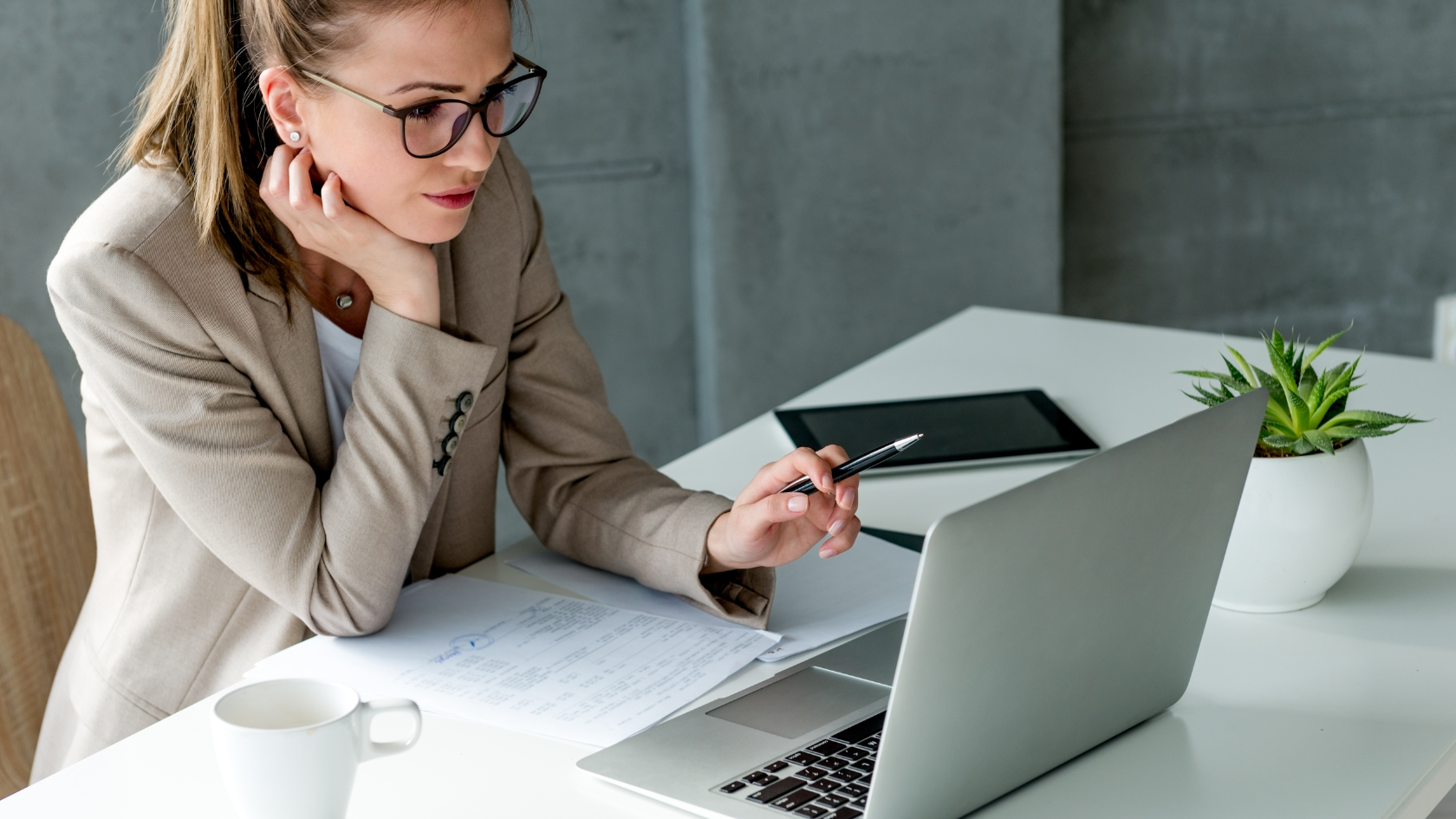 business woman holds a pen and rests her chin on her right hand while focusing on her laptop screen