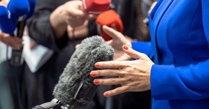 A woman dressed in a bright blue dress suit, speaks into a fuzzy mic head. There are other microphones being held up in the background as the woman in blue speaks.